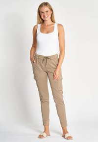 Beige Cargo-Hose mit elastischem Bund und Kordelzug. Verfügt über zulaufende Beine und Seitentaschen. Kombiniert mit einem enganliegenden weißen Tanktop.