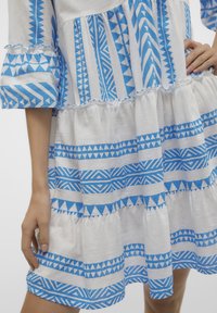 Robe blanche avec des motifs géométriques bleus, présentant des manches volantes et des niveaux, fabriquée en tissu léger avec une finition texturée.