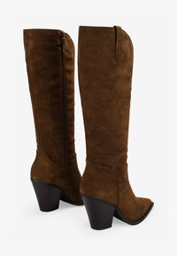 Botas de ante marrón hasta la rodilla con punta afilada, tacón alto apilado y cremallera lateral. Textura suave y diseño minimalista.