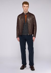 Chaqueta de cuero marrón con un alto cuello, combinada con un suéter gris con cremallera y pantalones azul marino. El modelo lleva mocasines marrón oscuro.