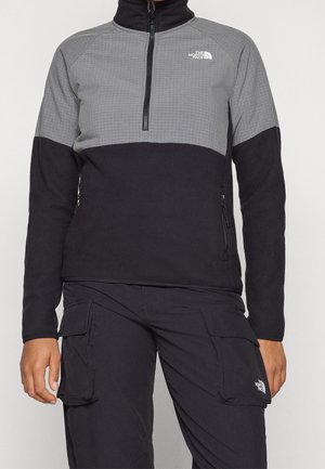 Person iført en sort og grå fleecejakke med kvart lynlås og sorte cargobukser, begge med The North Face-logo på brystet og benet.