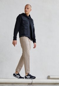 Svart flerfickad väst över en svart huvtröja, beige tapered byxor och svarta träningsskor med vita accenter, stående på en trappa.