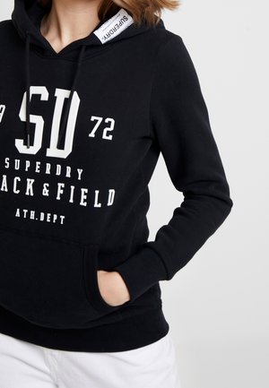 Personne portant un sweat à capuche noir Superdry avec texte et chiffres blancs, mains dans la poche avant, associé à un pantalon blanc sur un fond uni.