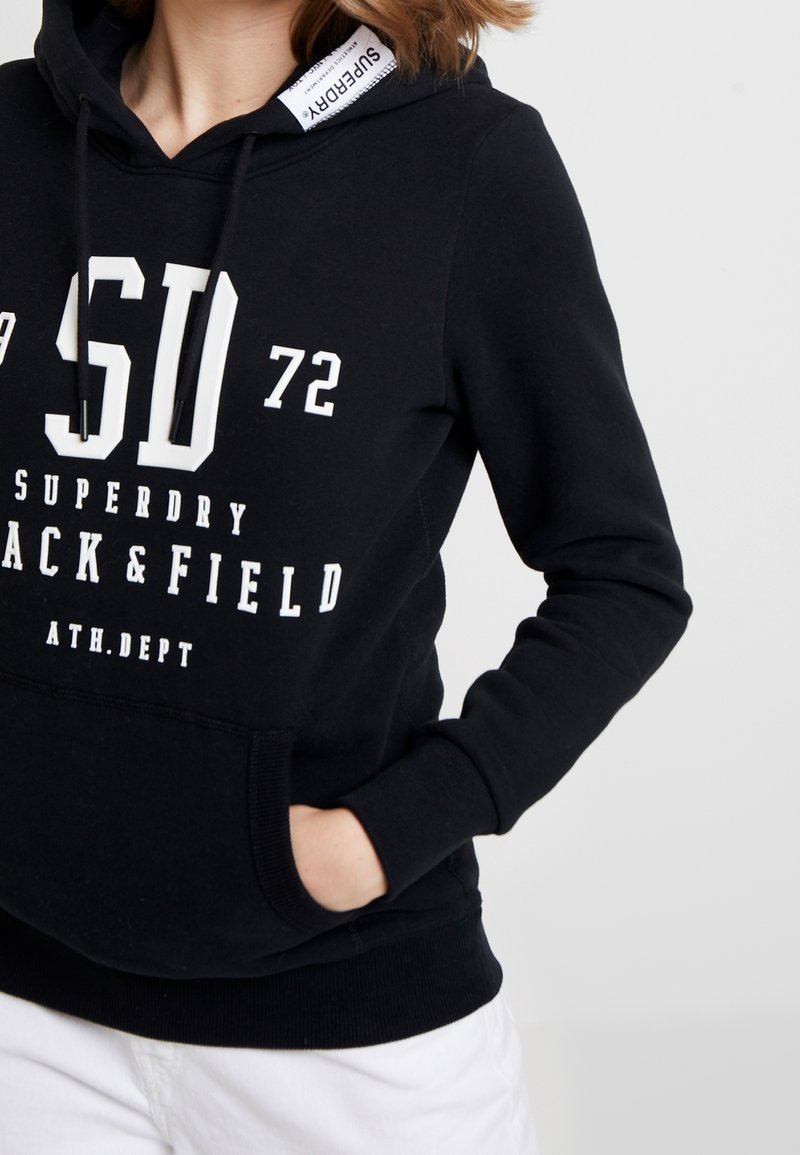 Personne portant un sweat à capuche noir Superdry avec texte et chiffres blancs, mains dans la poche avant, associé à un pantalon blanc sur un fond uni.