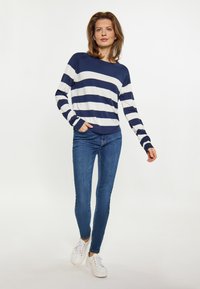 Marineblauwe en witte gestreepte katoenen trui met een ontspannen pasvorm. Gecombineerd met smalle blauwe jeans en witte sneakers. Het model staat zelfverzekerd.