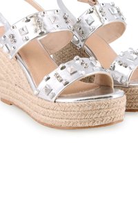 Sandalias de cuña plateadas con suela de yute trenzado, que presentan correas metálicas adornadas con tachuelas y gemas transparentes. Acabado suave y diseño abierto.