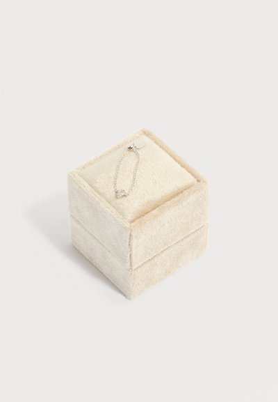 Μικρό ασημένιο βραχιόλι με μενταγιόν σε σχήμα καρδιάς, ακουμπισμένο σε ένα κλειστό μαλακό βελούδινο μπεζ κουτί κοσμημάτων πάνω σε λευκή επιφάνεια.