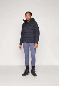 Czarna kurtka puchowa z wysokim kołnierzem i zamkiem błyskawicznym, zestawiona z jasnoszarymi legginsami i czarnymi butami trekkingowymi, tworząca warstwowy wygląd.