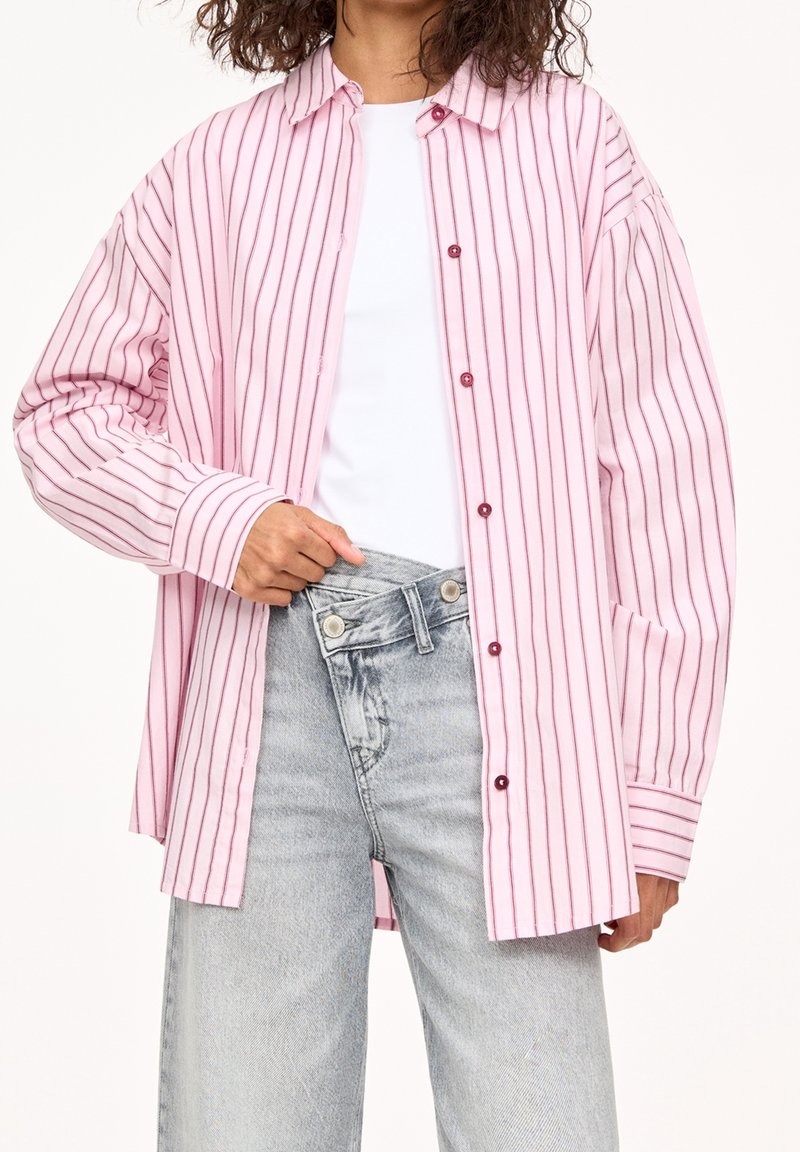 Femme portant une chemise à rayures verticales roses et blanches boutonnée par-dessus un haut blanc, associée à un jean taille haute gris clair, ajustant la ceinture.