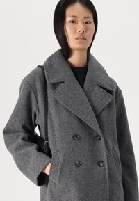 Cappotto grigio in lana doppiopetto con grandi revers, due bottoni neri e tasche laterali. Il modello tiene una borsa nera a tracolla.