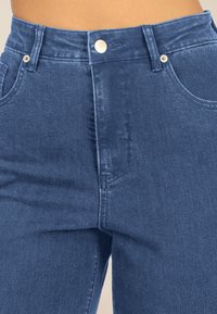 Denimjeans mit hoher Taille, einem Knopfverschluss und Vordertaschen. Der Stoff ist in einem mittleren Blauton gehalten, mit einer glatten Textur und einem leichten Glanz.
