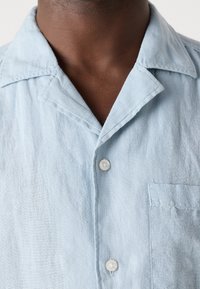 Gros plan du cou et du haut de la poitrine d'une personne portant une chemise en lin bleu clair à boutons, avec col et poche.