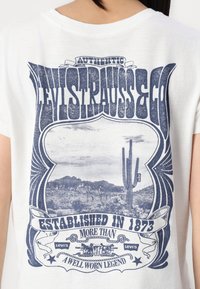 Balta kokvilnas T-krekls ar zilu grafiku, kurā attēloti kaktusi un kalni, ar uzrakstiem "Levi Strauss & Co" un "Izveidots 1873. gadā".