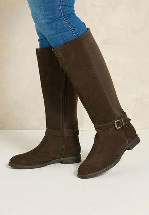 Bottes en daim marron montant jusqu'aux genoux avec fermeture éclair latérale et détail boucle, portées avec un jean skinny bleu sur un fond neutre.