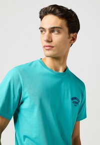 Turquoise katoenen t-shirt met een ronde hals, korte mouwen en een klein blauw geborduurd "Wrangler Jeans 1947" logo op de linker borst.