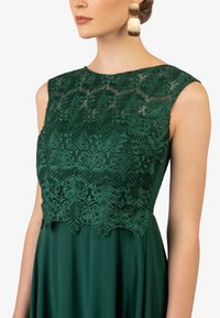 Smaragdgroen jurkje met een kanten lijfje met ingewikkelde bloempatronen, een ronde halslijn en vloeiend materiaal dat een elegante silhouet creëert.