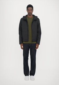 Schwarze gepolsterte Jacke mit Kapuze, getragen über einem olivgrünen Hemd und dunklen Jeans. Verfügt über einen Reißverschluss und elastische Bündchen. Weiße Sneakers runden das Outfit ab.