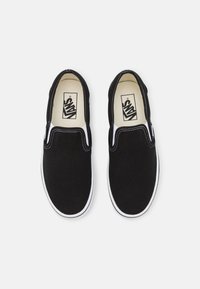Zapatillas de lona negras sin cordones con costuras blancas y suelas de goma. El forro interior es de color beige con un logo impreso en negro.