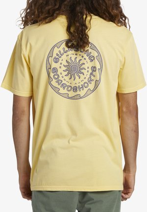 Camiseta de algodón amarilla con un gráfico circular de un sol en la parte posterior, con contornos negros y texto alrededor del diseño. Mangas cortas y corte holgado.