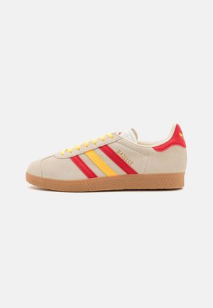 Beige suede sneaker with red and yellow stripes, featuring a rubber gum sole, flat yellow laces, and "Gazelle" logo in gold.