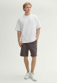 Camiseta blanca de gran tamaño combinada con pantalones cortos gris oscuro. El modelo lleva zapatillas blancas con detalles en marrón. Tejido de algodón suave con un ajuste relajado.