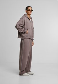 Felpa grigia oversize con tasca frontale e pantaloni a sigaretta coordinati, caratterizzati da strisce laterali bianche e tessuto texturizzato. Le sneakers completano il look.