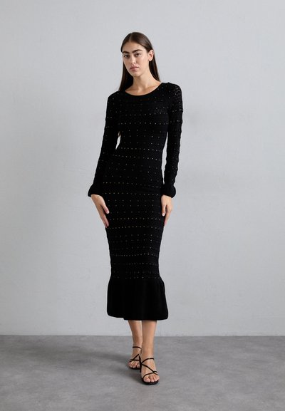 Woman wearing a black, long-sleeve, ankle-length fitted dress with small studs and ruffle hem, paired with black strappy sandals.
