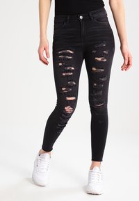 Jeans skinny negros con múltiples áreas desgastadas, con bordes deshilachados. Combinados con zapatillas blancas, enfatizando el diseño desgastado.