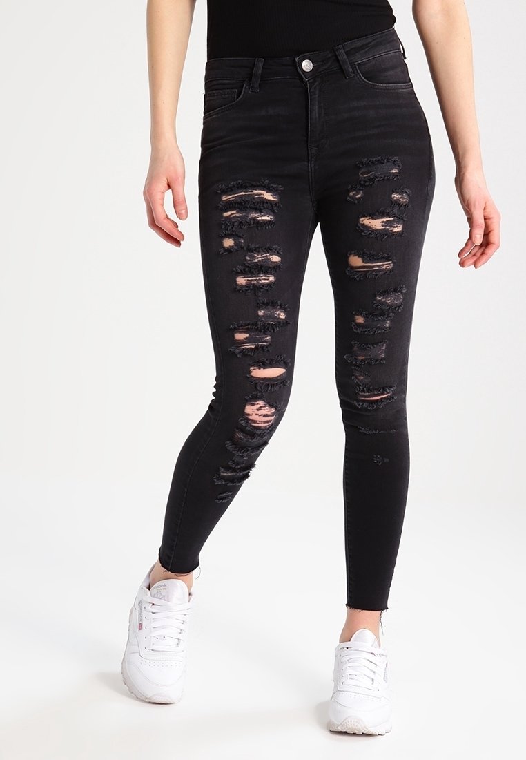 Jeans skinny negros con múltiples áreas desgastadas, con bordes deshilachados. Combinados con zapatillas blancas, enfatizando el diseño desgastado.
