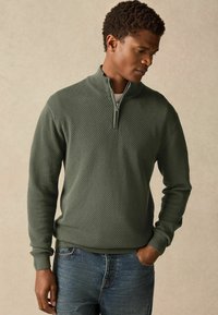 Dunkelgrüner Zip-Kragen-Pullover mit strukturiertem Muster, gerippten Bündchen und Saum. Wird mit engen blauen Jeans getragen.