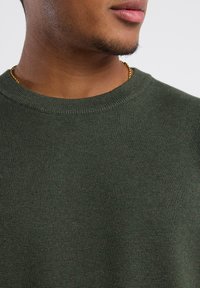 Dark green knit sweater with a crew neckline and ribbed texture. Features a close-up view of the fabric and neckline details.