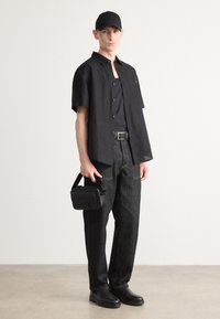 Camisa negra de mangas cortas, top negro sin mangas, pantalones de mezclilla oscuros, cinturón negro, zapatos negros y bolsa negra pequeña, con un fondo neutro.