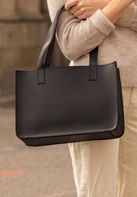 Sac fourre-tout en cuir noir avec une texture lisse, de forme rectangulaire et deux poignées robustes. Présente un design minimaliste et un logo en relief.