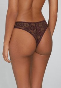 Burgunderfarbene Spitzenstringtanga mit floralen Mustern, mit transparentem Design und gewellten Kanten, die das strukturierte Detail betonen.