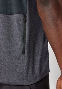 T-shirt atlético cinza escuro com um bolso com fecho, tecido texturizado e detalhe de logótipo discreto perto da parte inferior. Apresenta um painel contrastante na parte superior.