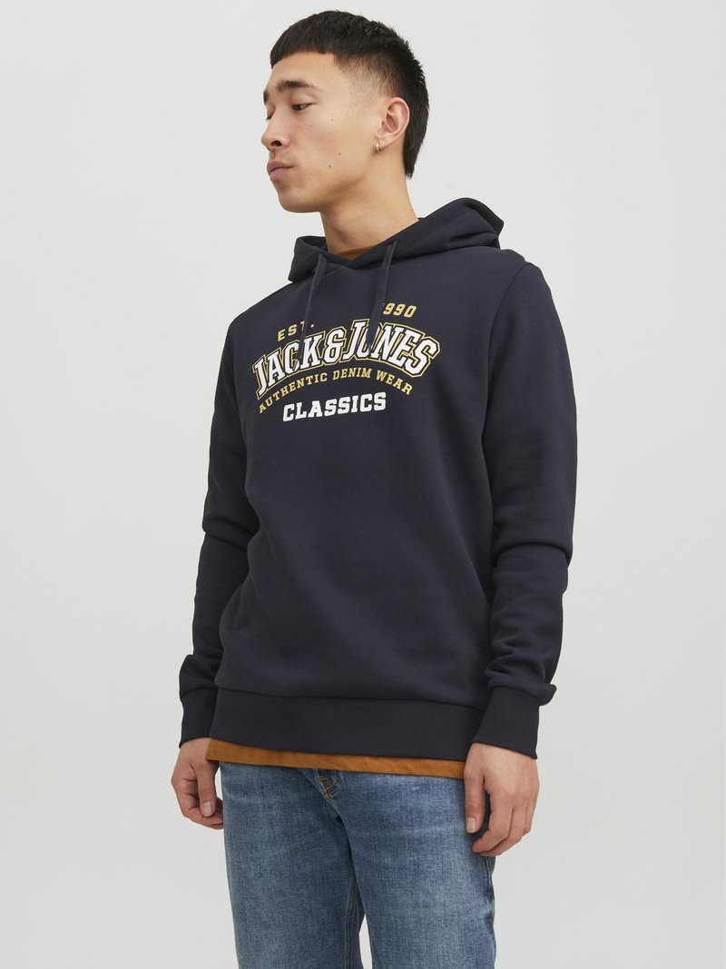Felpa navy con grafica davanti, cappuccio con cordoncino, tasca a marsupio e polsini a coste. Abbinata a una maglietta beige e jeans azzurri chiari.