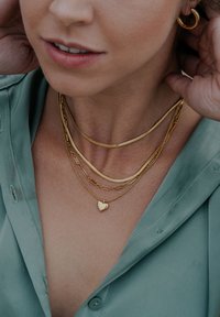 Coliere din aur cu straturi și modele variate: un lanț subțire, zale decorative și un pandantiv mic în formă de inimă, purtate peste o cămașă verde deschis.