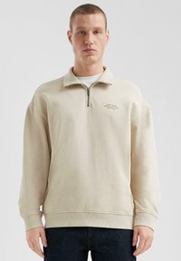 Felpa leggera beige con zip e colletto alto, maniche lunghe, polsini a coste e orlo dritto. Presenta testo ricamato sul petto sinistro.