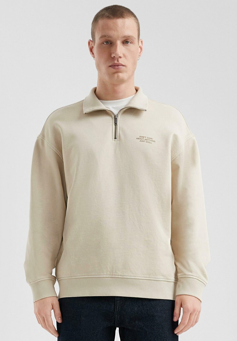 Felpa leggera beige con zip e colletto alto, maniche lunghe, polsini a coste e orlo dritto. Presenta testo ricamato sul petto sinistro.