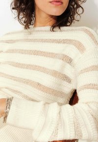 Pull en tricot rayé blanc et beige avec un motif texturé, des manches larges et des détails en tricot ouvert, drapé sur une chaise en bois.