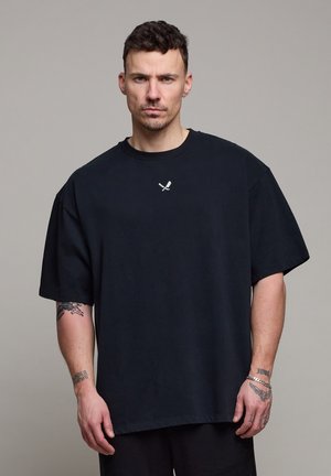 Mann med kort krøllete hår som har på seg en oversized svart T-skjorte med en liten logo av kryssete kniv og gaffel, og viser tatoveringer på armer og hender.