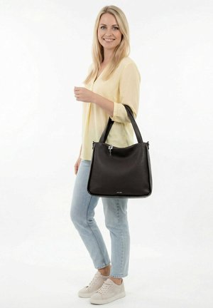 Blonde woman wearing light yellow blouse and jeans, smiling, holding a large black leather handbag over her shoulder.