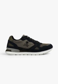 Zapatilla negra y oliva oscuro con logo de calavera y huesos cruzados en el lateral, suela blanca y cierre con cordones, vista desde el lado interior.
