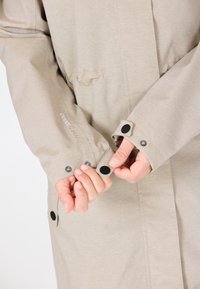 Manteau de pluie beige avec des manches longues, des poignets réglables dotés de boutons noirs, et un tissu texturé subtil ; logo de la marque visible sur la manche.