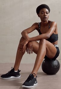 Sort sports-bh og shorts båret med sorte træningssko. Sidder på en sort medicinalbold, glatte teksturer, minimalistisk design, neutral baggrund.