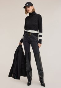 Czarny sweter z białymi paskami, prążkowany golf, zestawiony z czarnymi, wysokim stanem jeansami i błyszczącymi czarnymi kozakiem za kolano. Trzyma kurtkę.