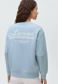 Maglione azzurro chiaro con collo rotondo, caratterizzato da una grafica testuale bianca testurizzata sul retro. Design casual con vestibilità comoda e polsini a coste.