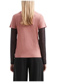 Blond inimene, kes kannab tolmuroosa lühikeste varrukatega särki, mille all on musta läbipaistva pikkade varrukatega särk, millel on leopardimuster, koos tumedate triibuliste pükstega.