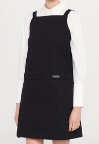 Vestido negro sin mangas con escote cuadrado, tirantes ajustables y un sutil detalle del logo de LPS en la cintura, combinado con una camisa blanca con cuello.