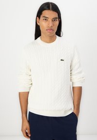 Hvit kabelstrikket genser med ribbestrikkede mansjetter og kant, med en liten grønn brodert logo på brystet. Glatt tekstur.